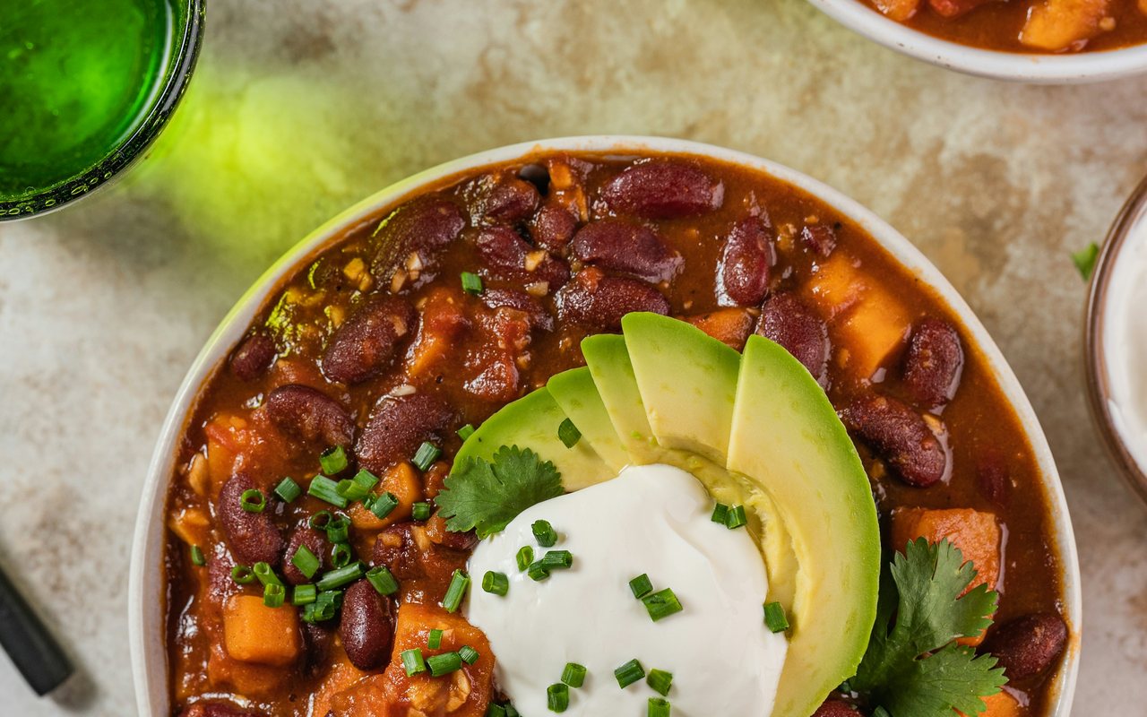 A bowl of red bean and sweet potato chili topped with avocado, cilantro, chives, and a spoonful of white yogurt.