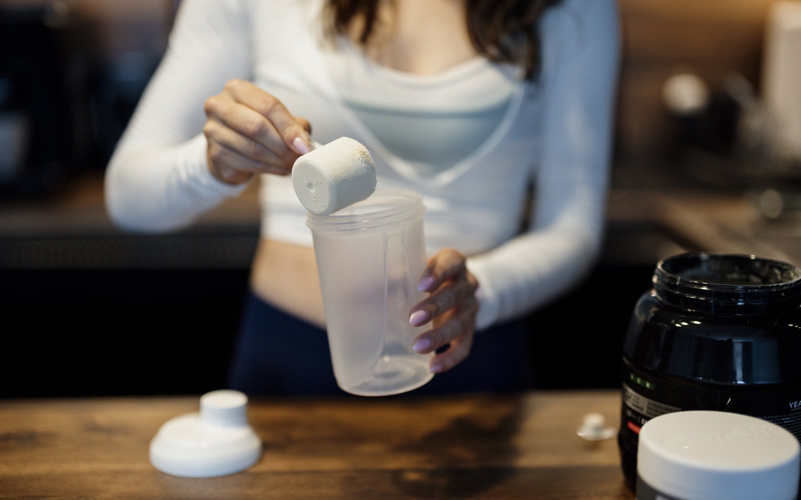 Woman scoops white powder into a shaker bottle beside an open supplement tub.