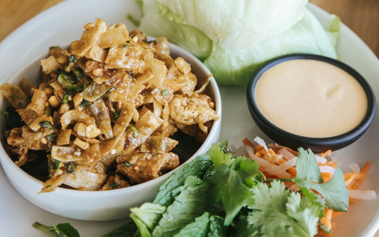 A bowl of chopped meat and vegetables beside iceberg lettuce, herbs, shredded slaw, and a cup of pale dipping sauce.