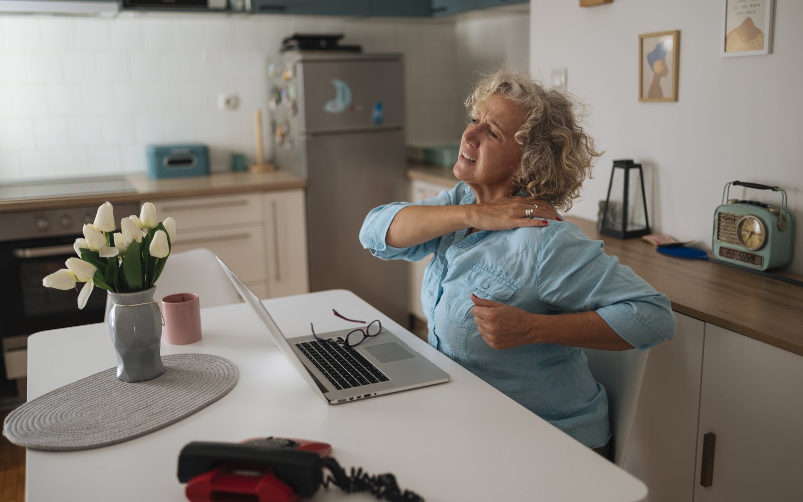 Older adult seated at a laptop touches one shoulder in a home workspace.