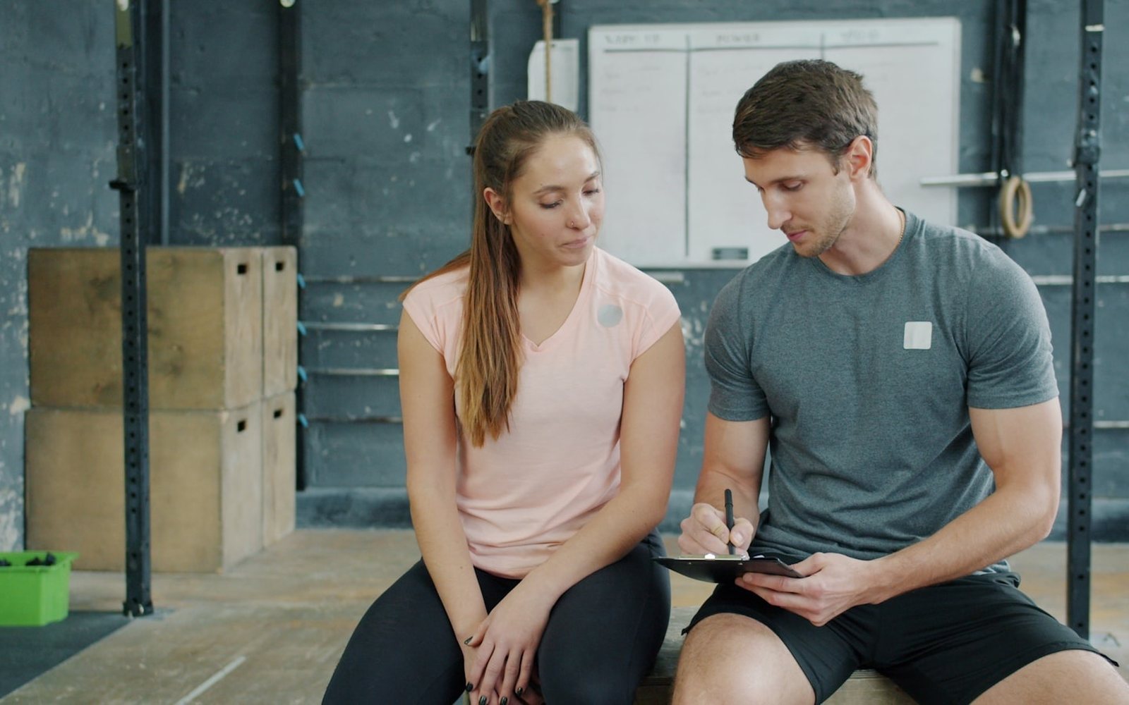 Trainer writes on a clipboard while seated beside a gym client.