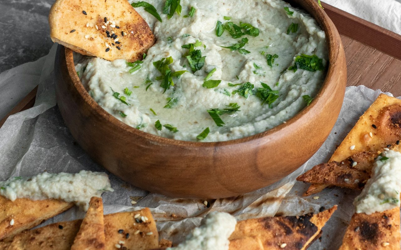 A wooden bowl of pale green eggplant dip topped with chopped herbs and surrounded by toasted pita chips.