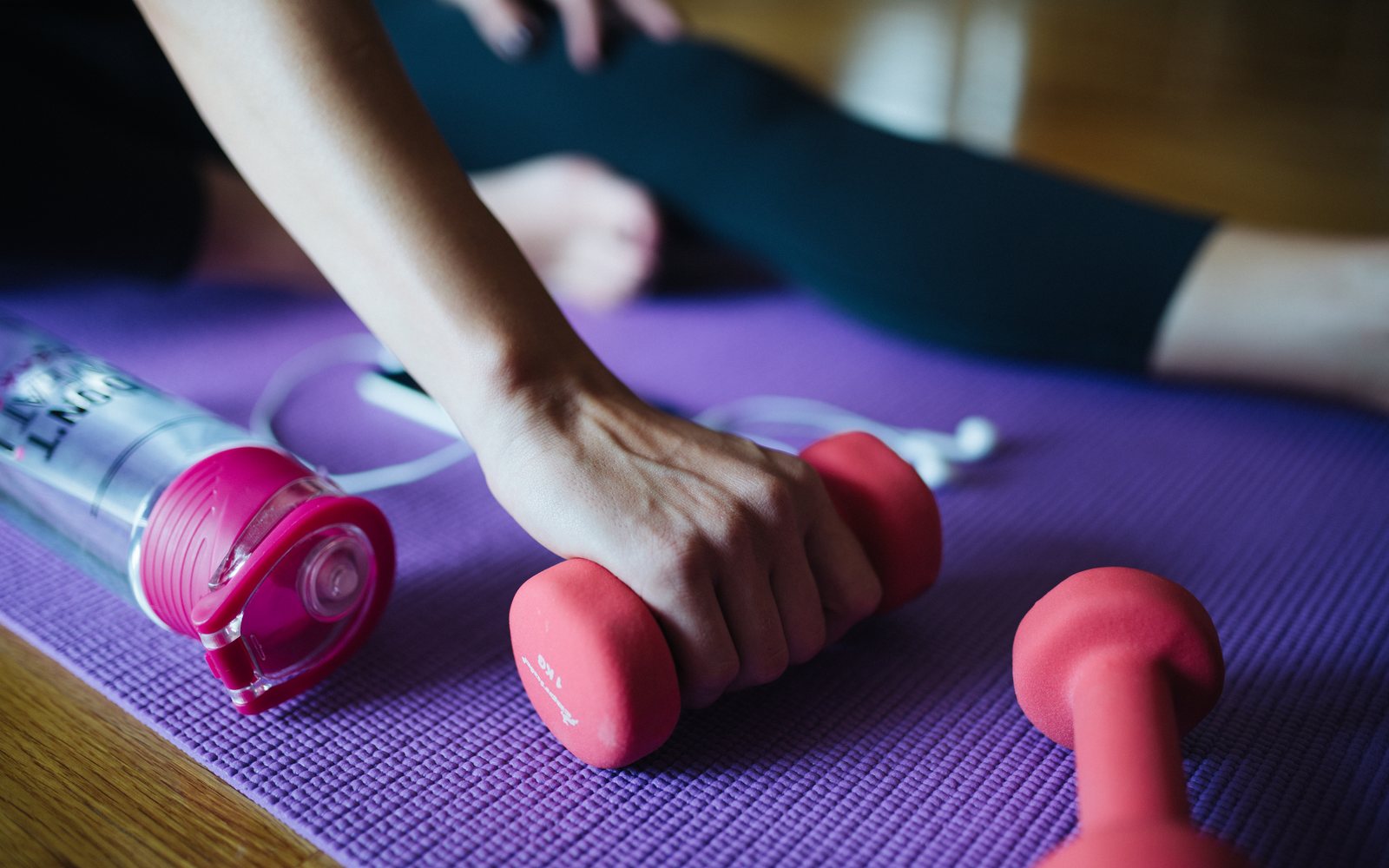 Hand grips a pink dumbbell on a purple mat beside another dumbbell and a water bottle.