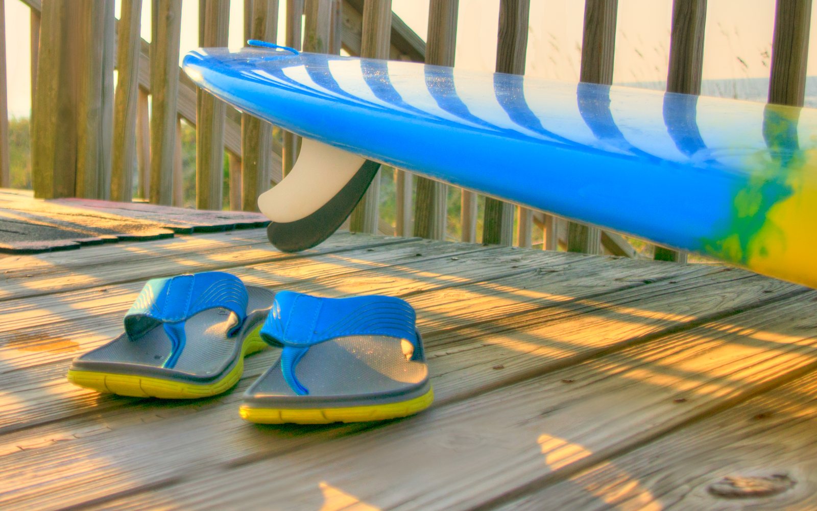 Blue flip-flops rest beside a blue surfboard on a sunlit wooden deck.
