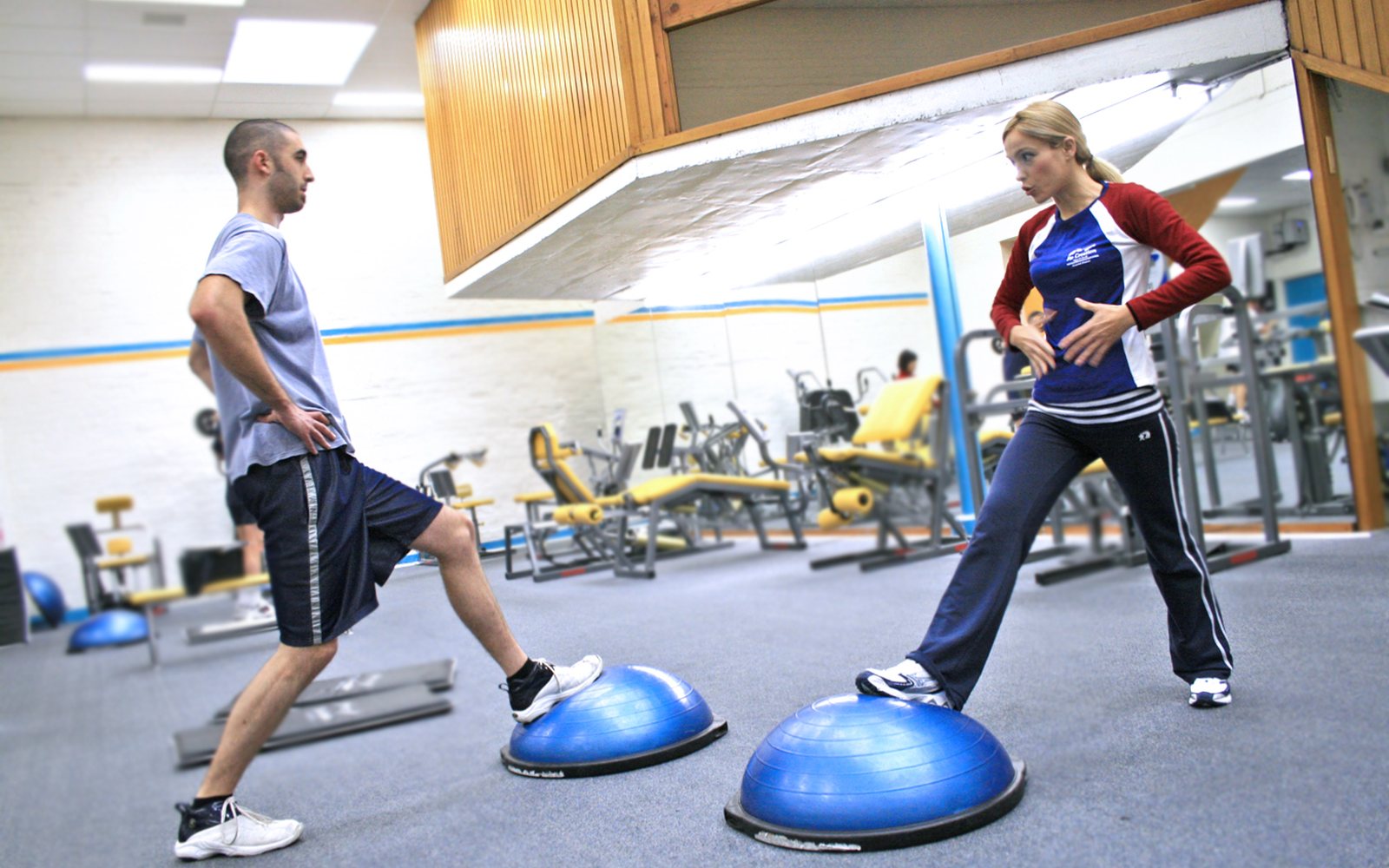 Man and woman each balance one foot on a blue half-dome trainer in a gym.