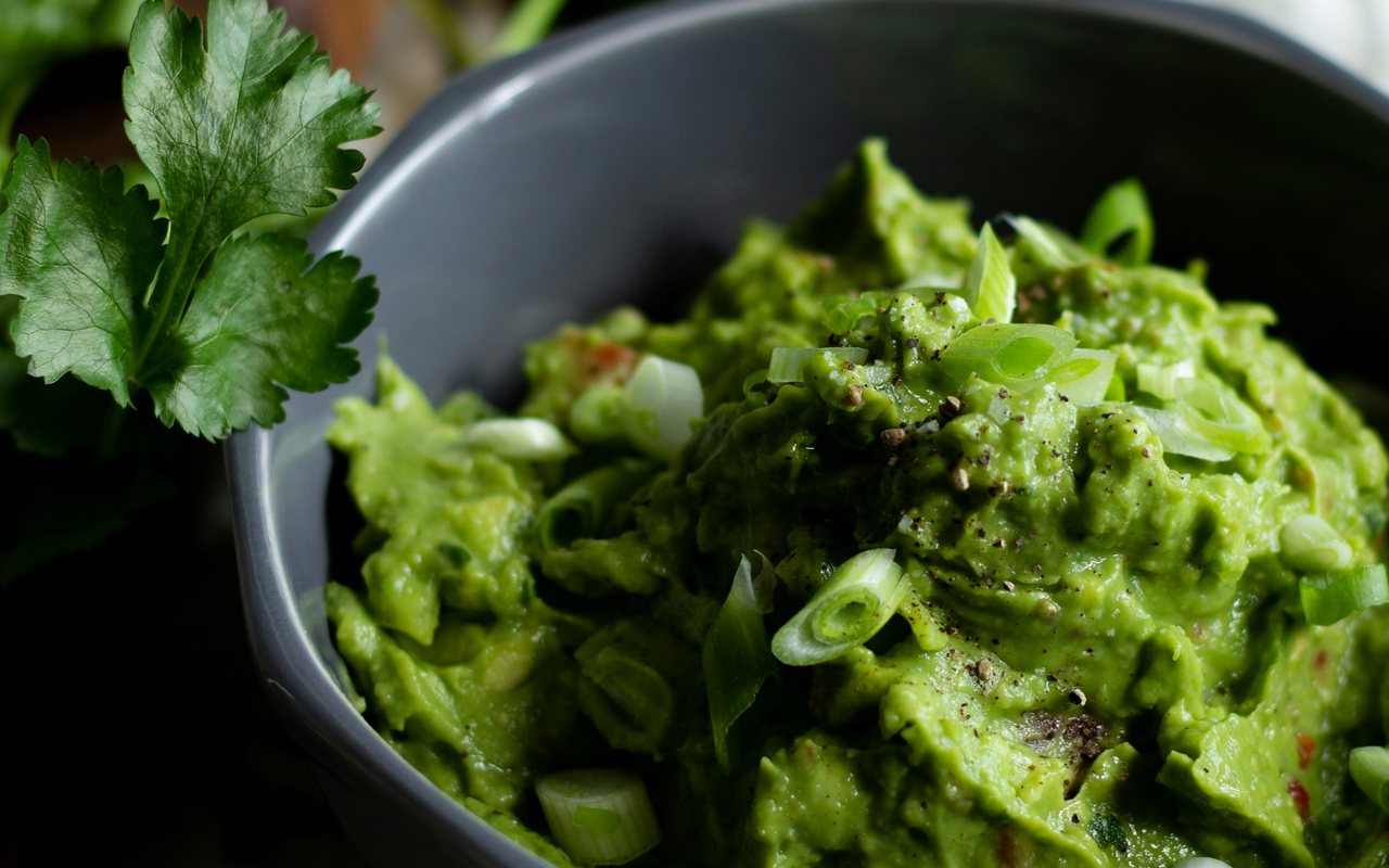 A dark gray bowl of chunky green avocado dip topped with sliced scallions and cracked pepper beside fresh cilantro.