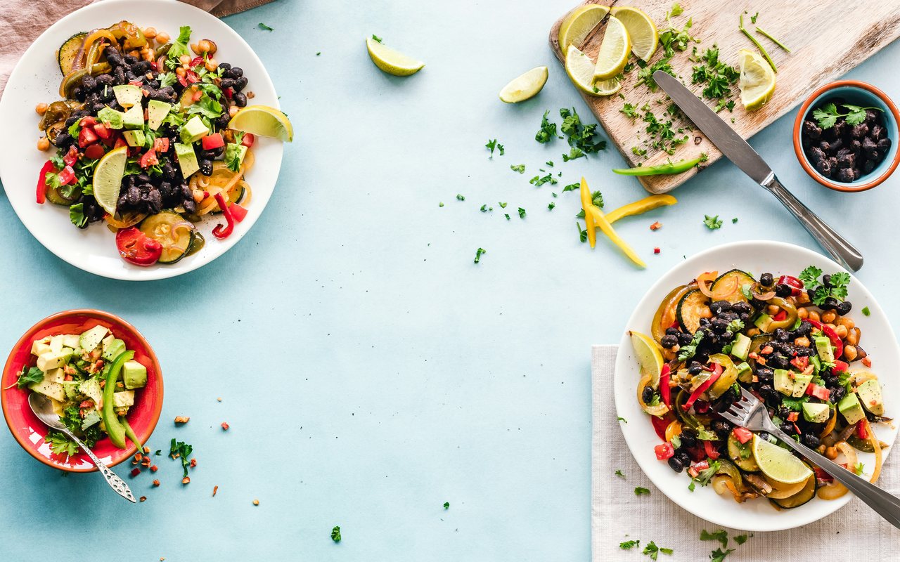 Three bowls of black beans and colorful fajita vegetables with avocado and lime on a pale blue table.