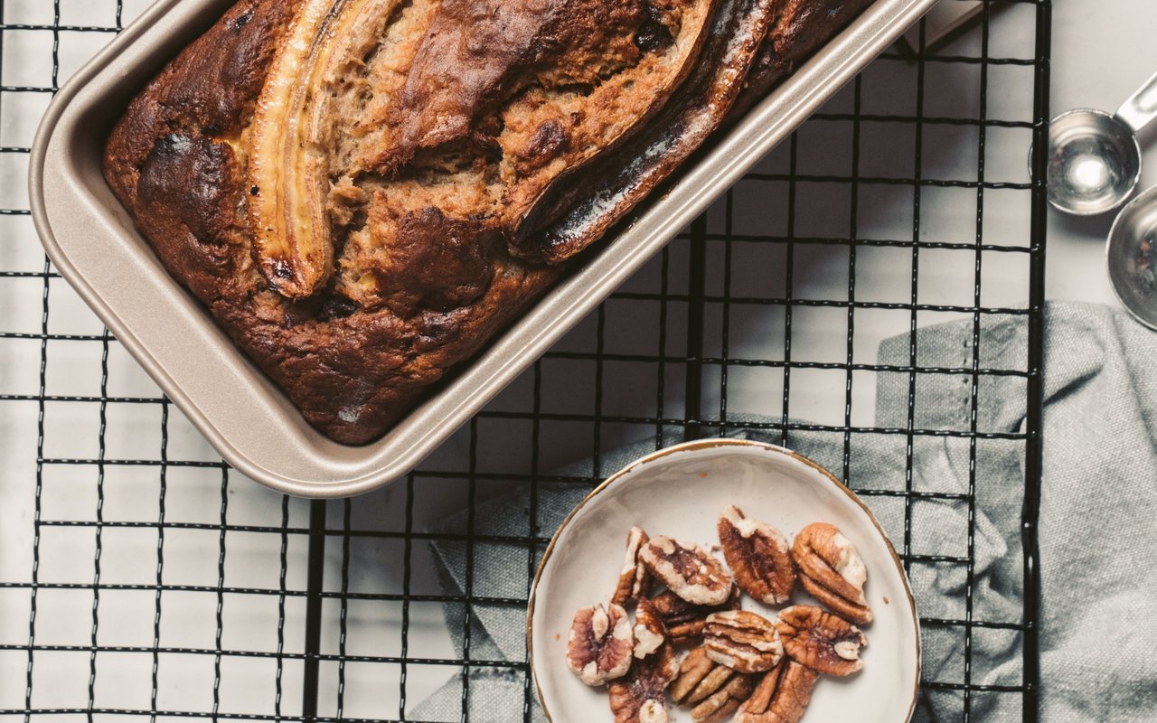Dark banana bread loaf topped with baked banana halves beside pecans