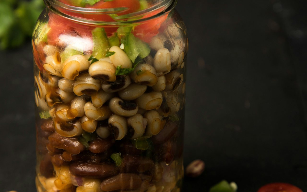 A glass jar layered with mixed beans, cucumber, tomato, and chopped herbs.
