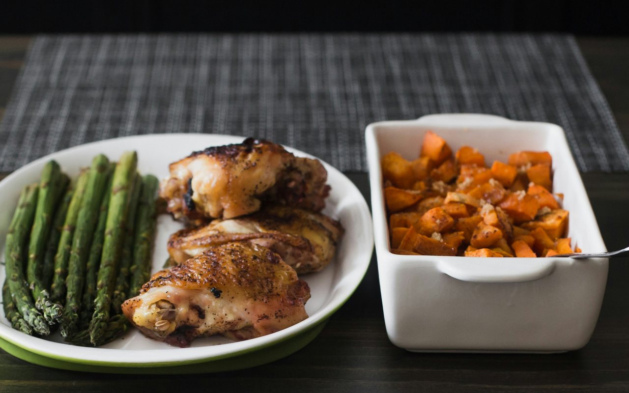 Roasted chicken thighs with asparagus and cubed sweet potatoes in white serving dishes.