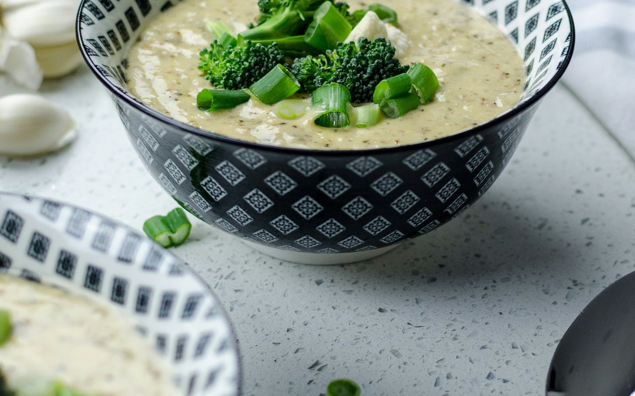 Creamy broccoli and cauliflower soup topped with broccoli florets and scallions in a patterned bowl.