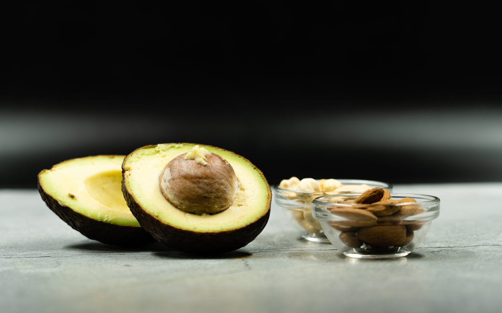 Halved avocado sits beside small glass bowls of almonds and cashews.