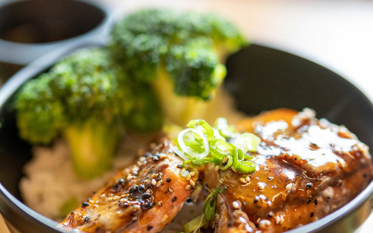 Glazed salmon over rice with broccoli and sliced green onions in a black bowl.