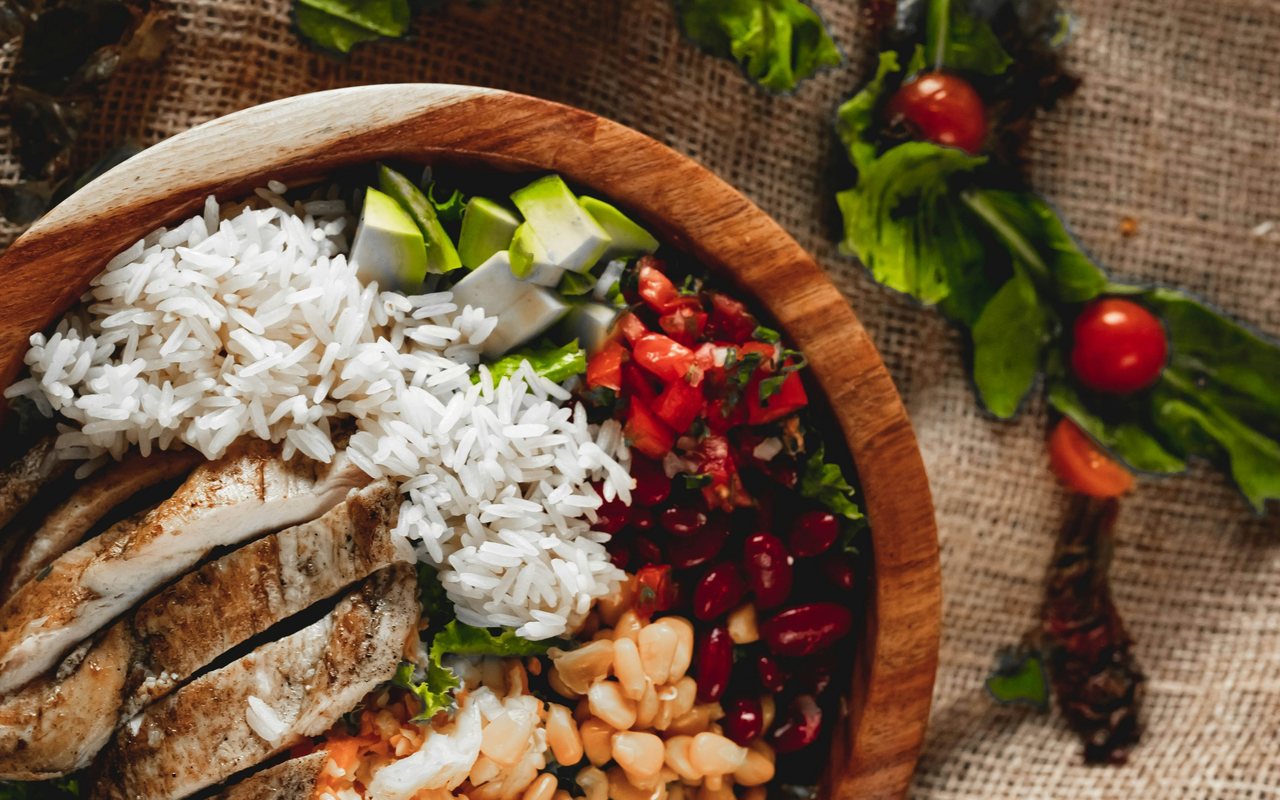 A wooden bowl with sliced grilled chicken, white rice, kidney beans, corn, avocado, chopped tomato, and lettuce.