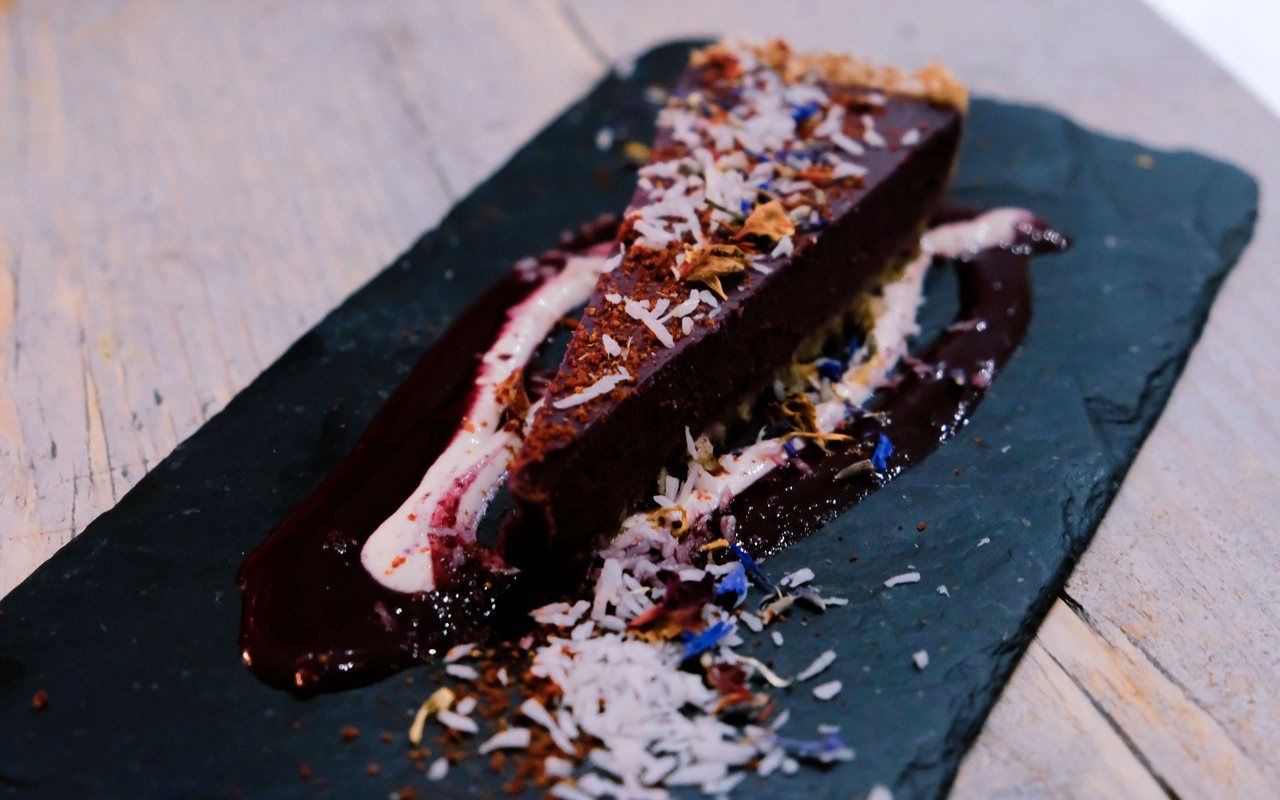 Dark chocolate cheesecake slice with chocolate sauce and coconut flakes on a black slate plate