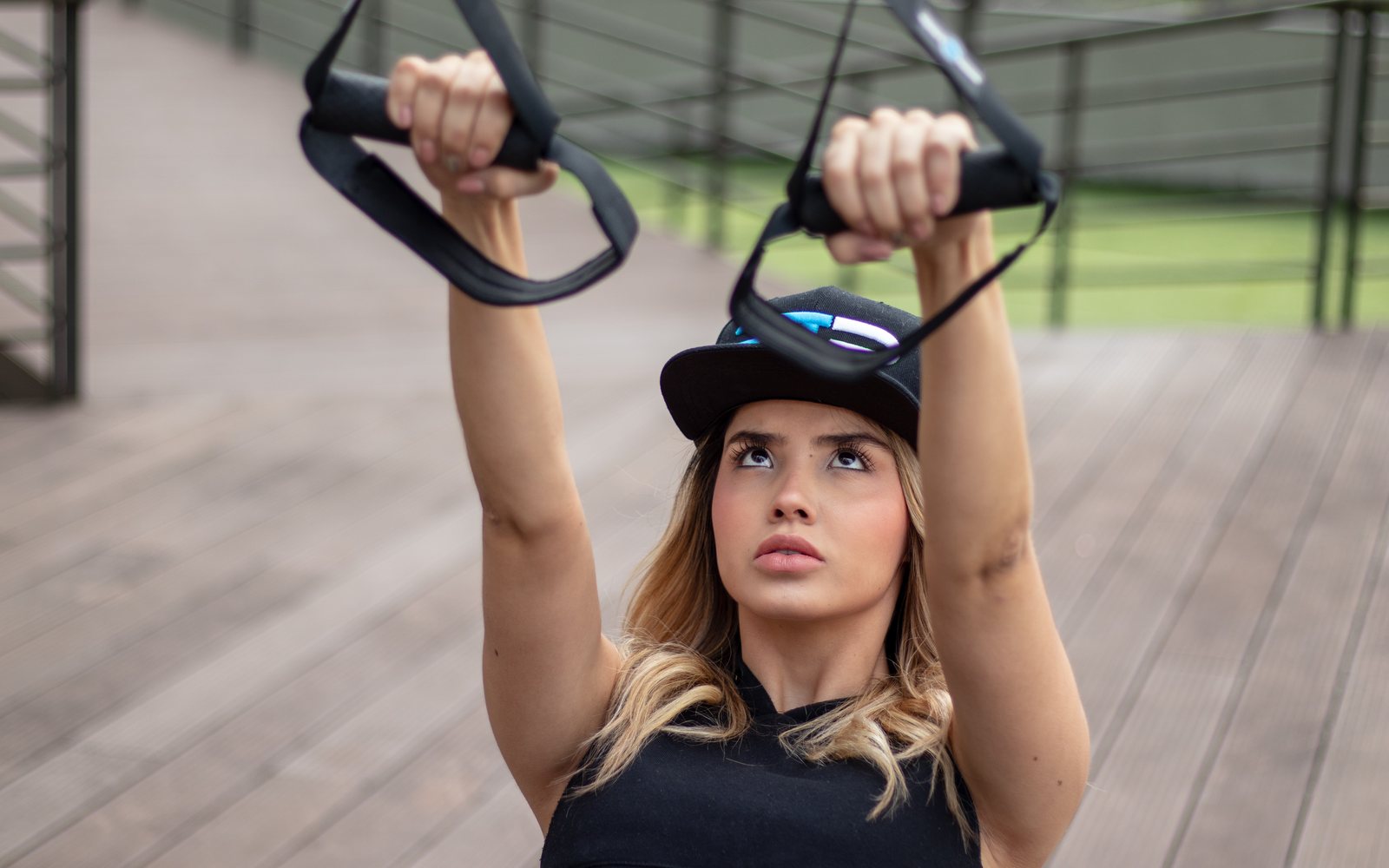 Woman grips suspended exercise handles overhead on an outdoor deck.