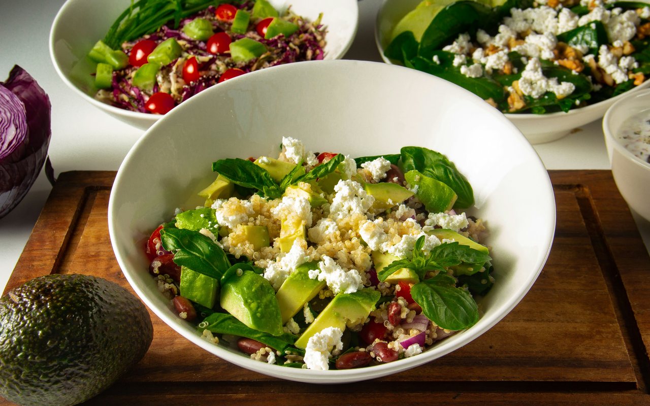 Three white bowls of leafy salad with avocado, cherry tomatoes, crumbled feta, quinoa, and herbs on a wooden board.