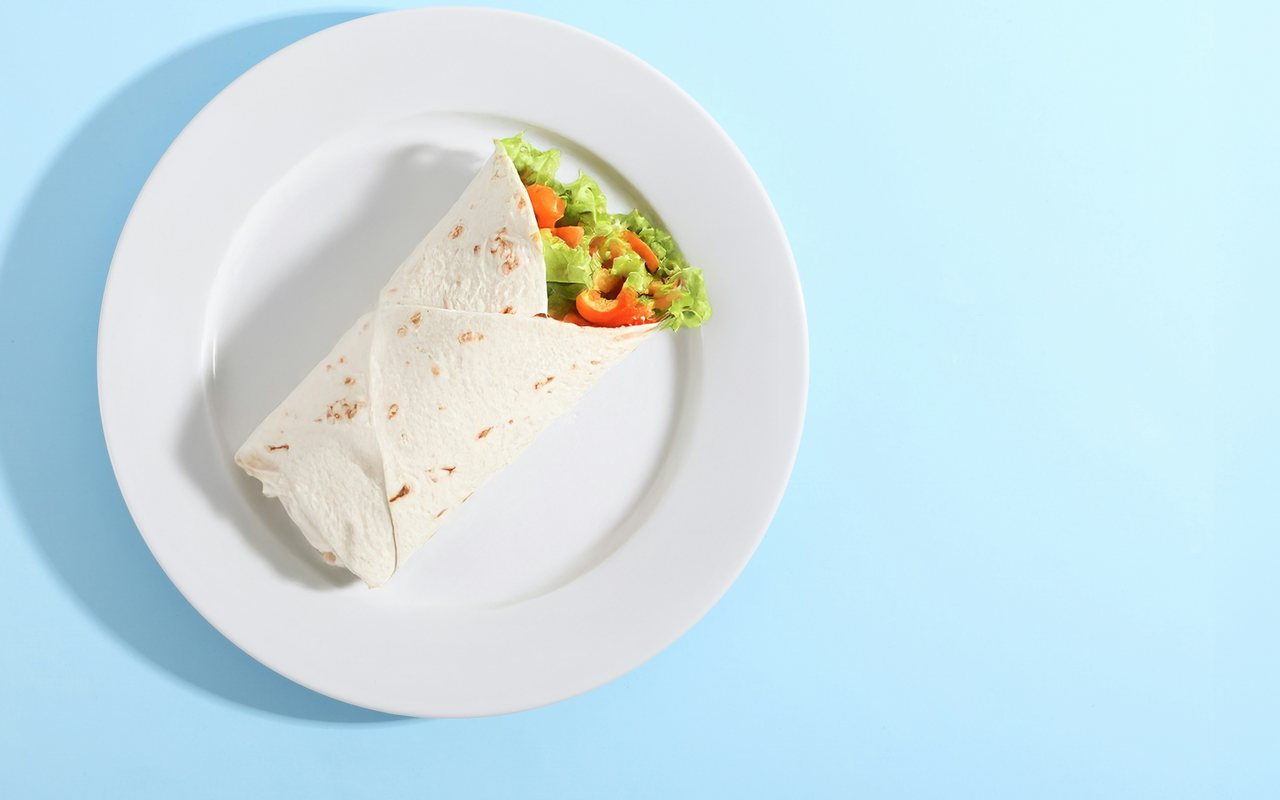 A tortilla wrap filled with lettuce and sliced red and yellow peppers on a white plate against a blue background.