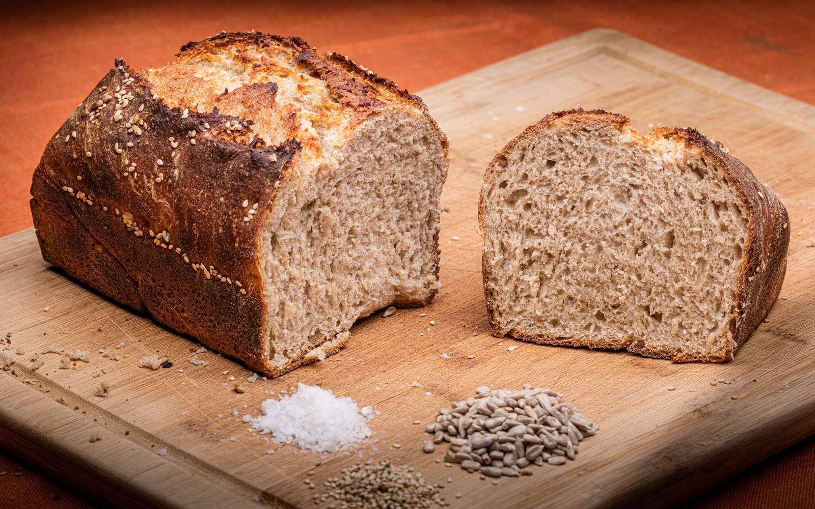 Cut loaf and slice of seeded bread rest on a wooden cutting board.