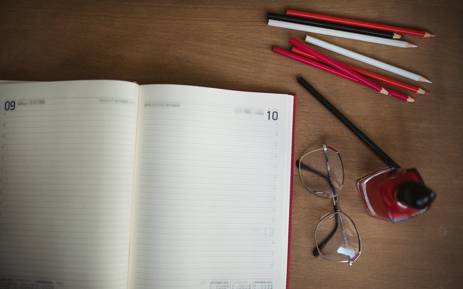 Open dated planner beside colored pencils, eyeglasses, and red nail polish on a wooden desk.