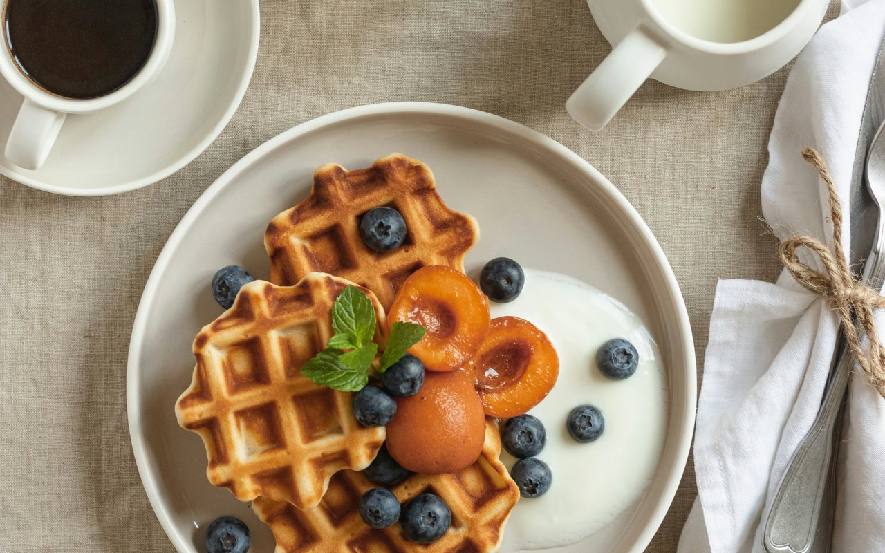 Mini waffles with blueberries, apricot halves, and a spoonful of white yogurt on a breakfast table.