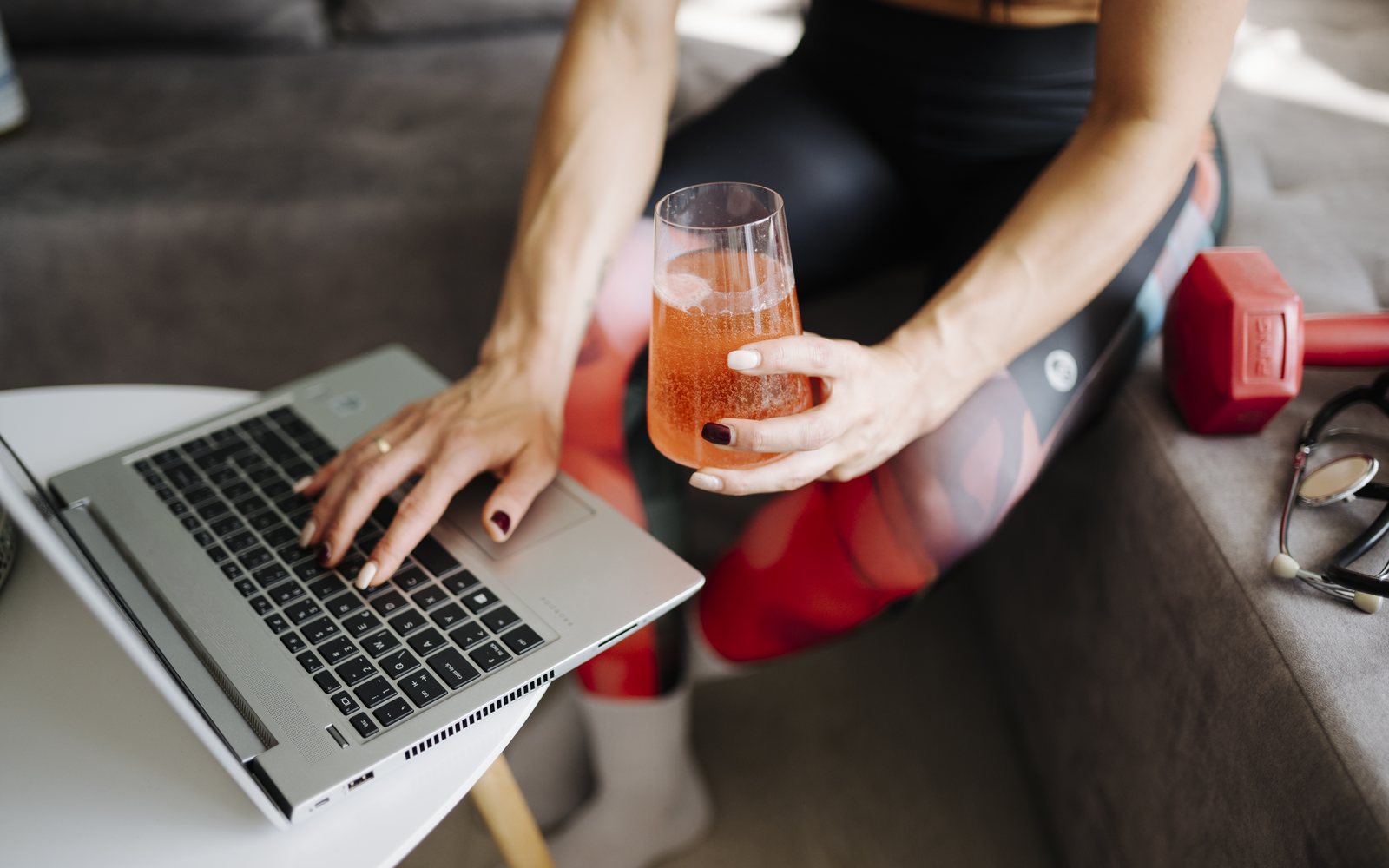Adult in workout clothes reviews a laptop beside a dumbbell while holding an orange drink.