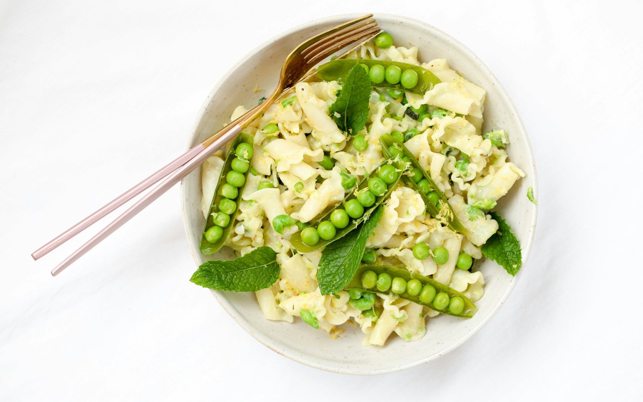 A white bowl of ruffled lemon pasta with green peas and fresh mint on a white table.