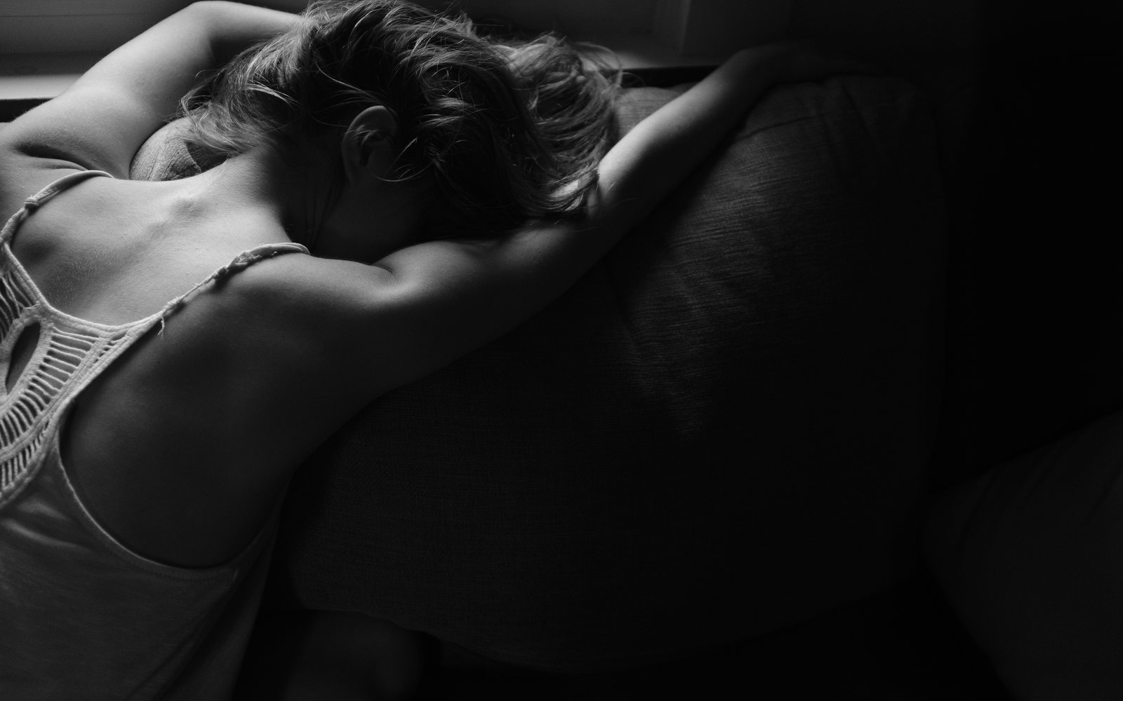 Person rests with their head on folded arms on a dark sofa in black and white.