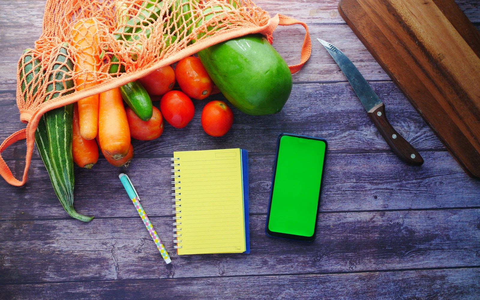 Fresh vegetables, a notebook, and a smartphone arranged on a kitchen table
