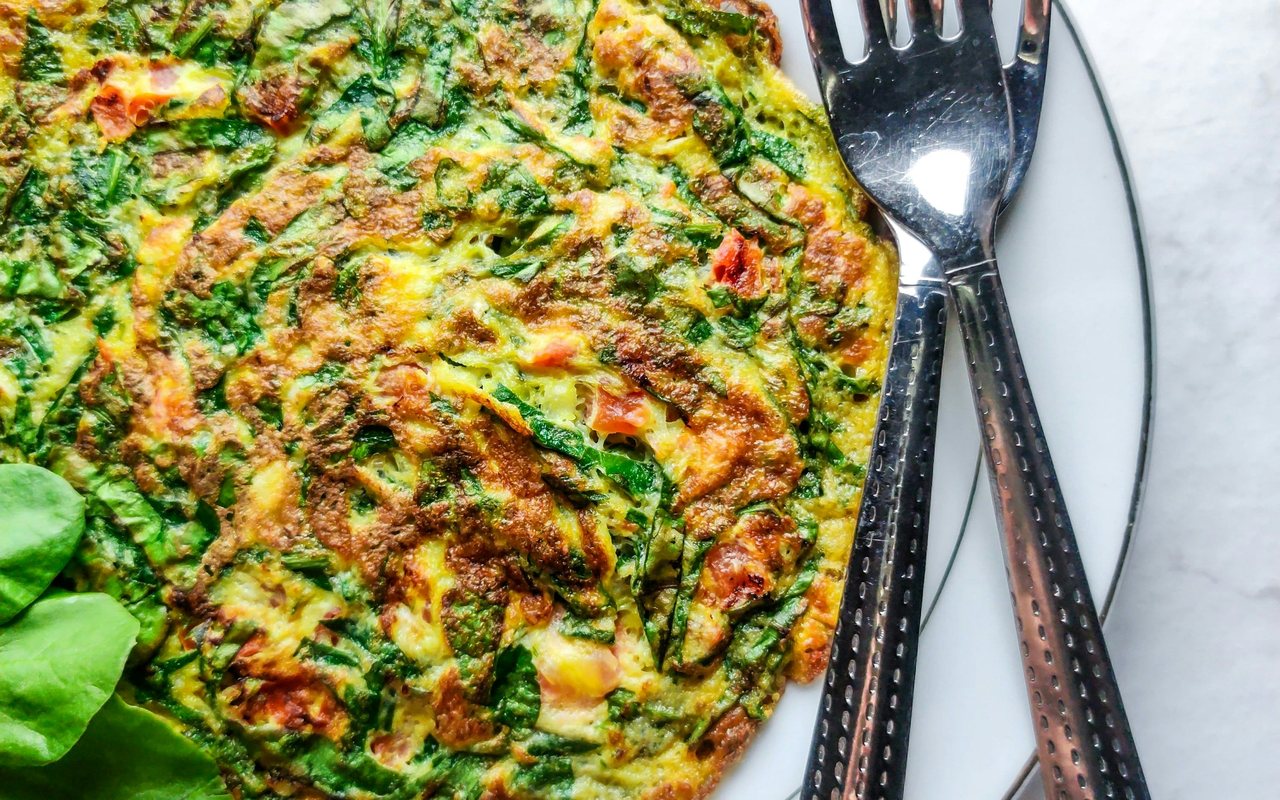 A browned spinach and tomato omelet on a white plate beside two metal forks.