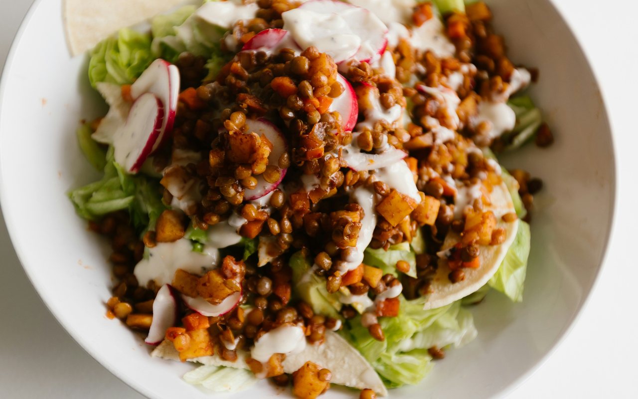 Lettuce and lentil salad with radishes, diced vegetables, tortilla pieces, and creamy white dressing in a white bowl.