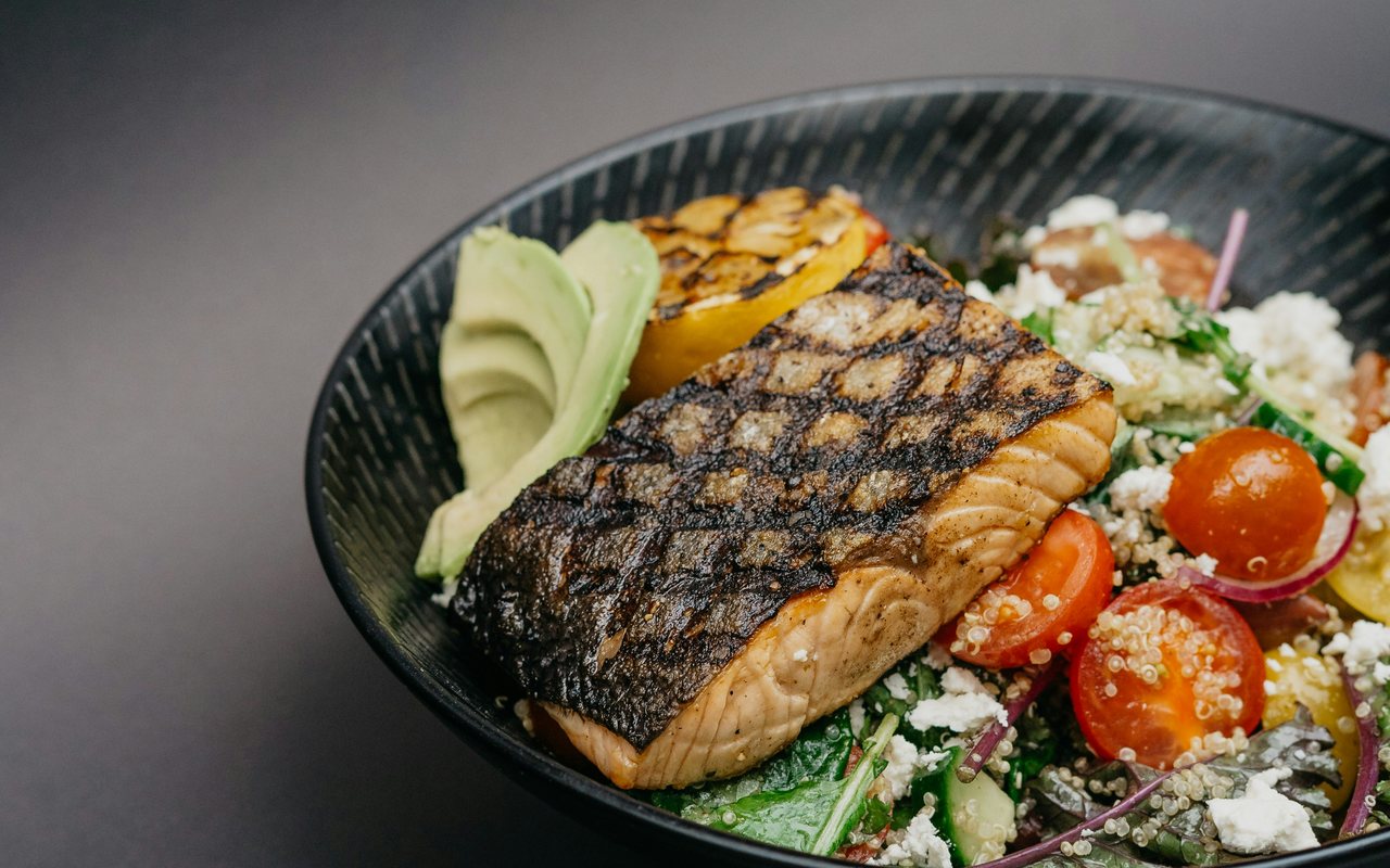 A charred salmon fillet with avocado, quinoa, leafy greens, cherry tomatoes, red onion, crumbled cheese, and grilled yellow pepper in a black bowl.