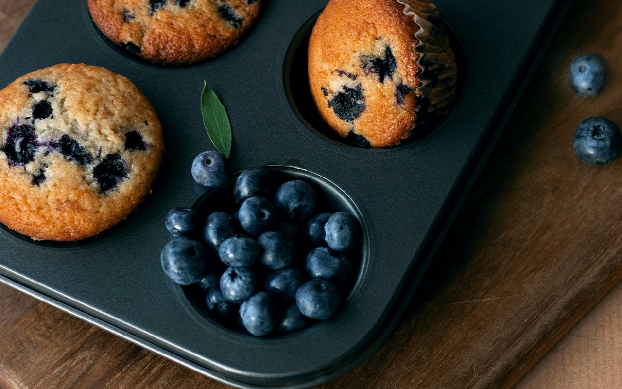 Blueberry baked cups in a dark muffin tin beside a bowl of fresh blueberries.