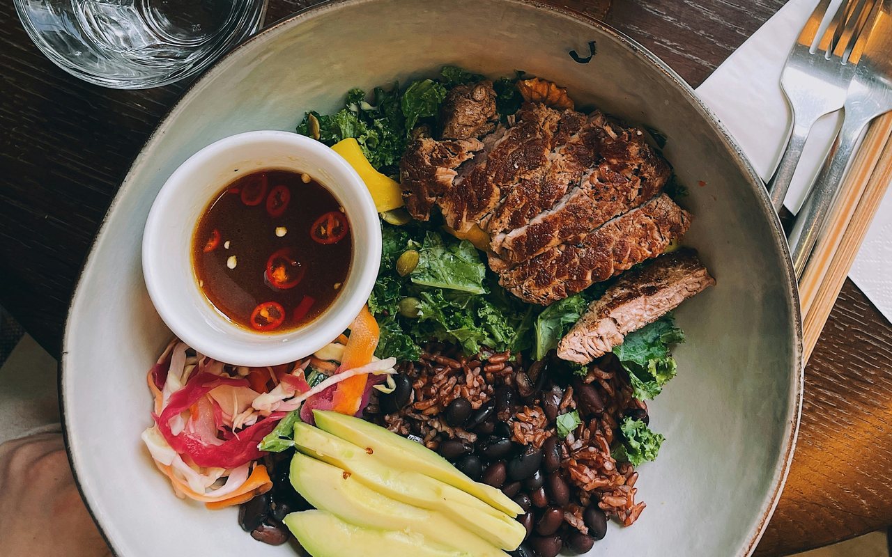Sliced steak with red rice, black beans, avocado, kale, slaw, and a bowl of chile sauce.