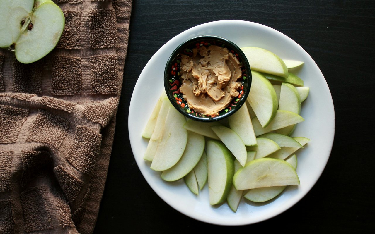 Green apple slices arranged around a small bowl of peanut butter on a white plate.
