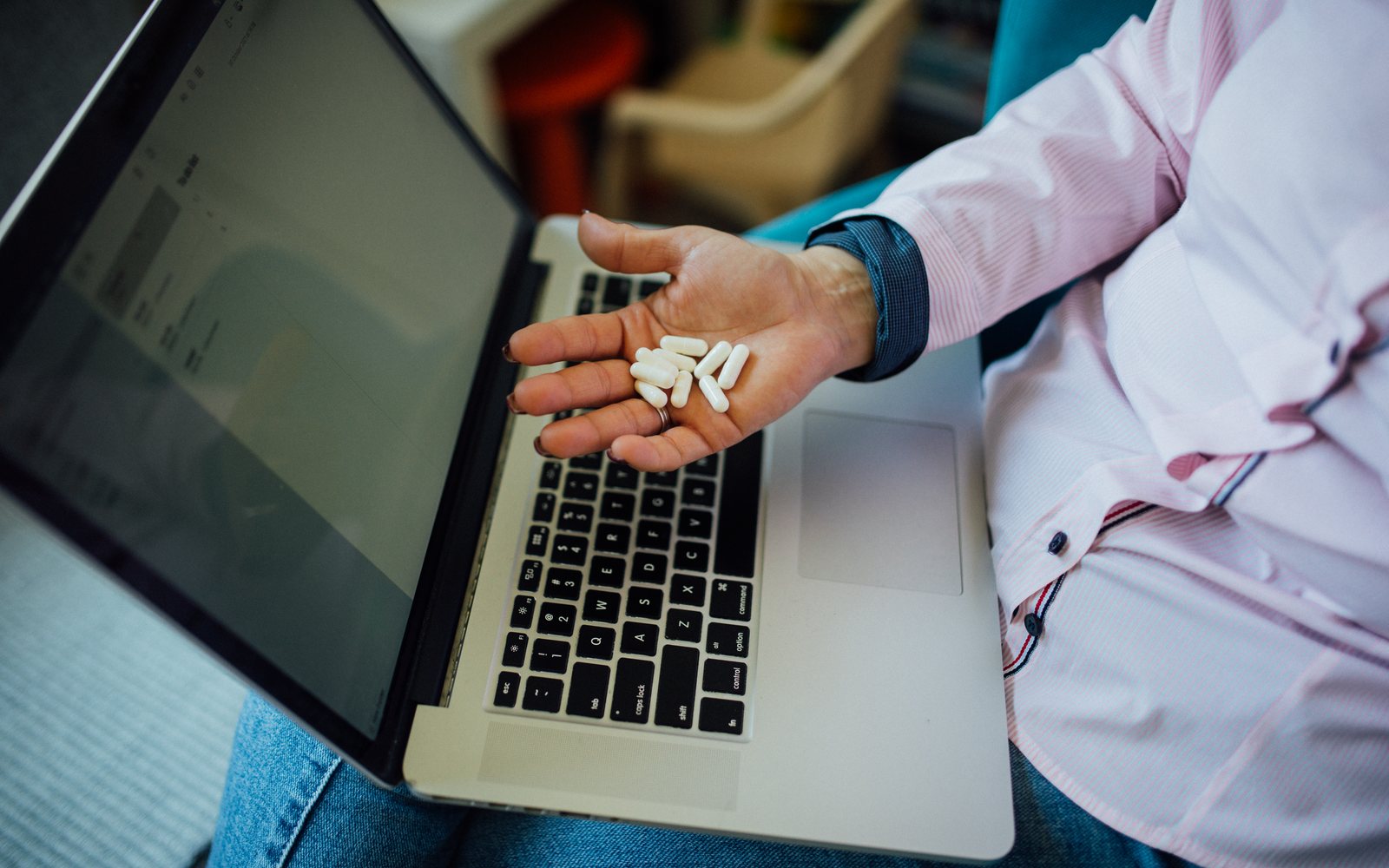 Adult holds several white capsules above an open laptop while researching supplements.
