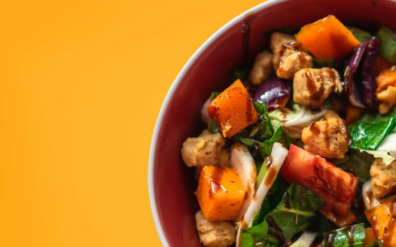 A red bowl of browned protein pieces, roasted orange squash, tomatoes, cabbage, and leafy greens.
