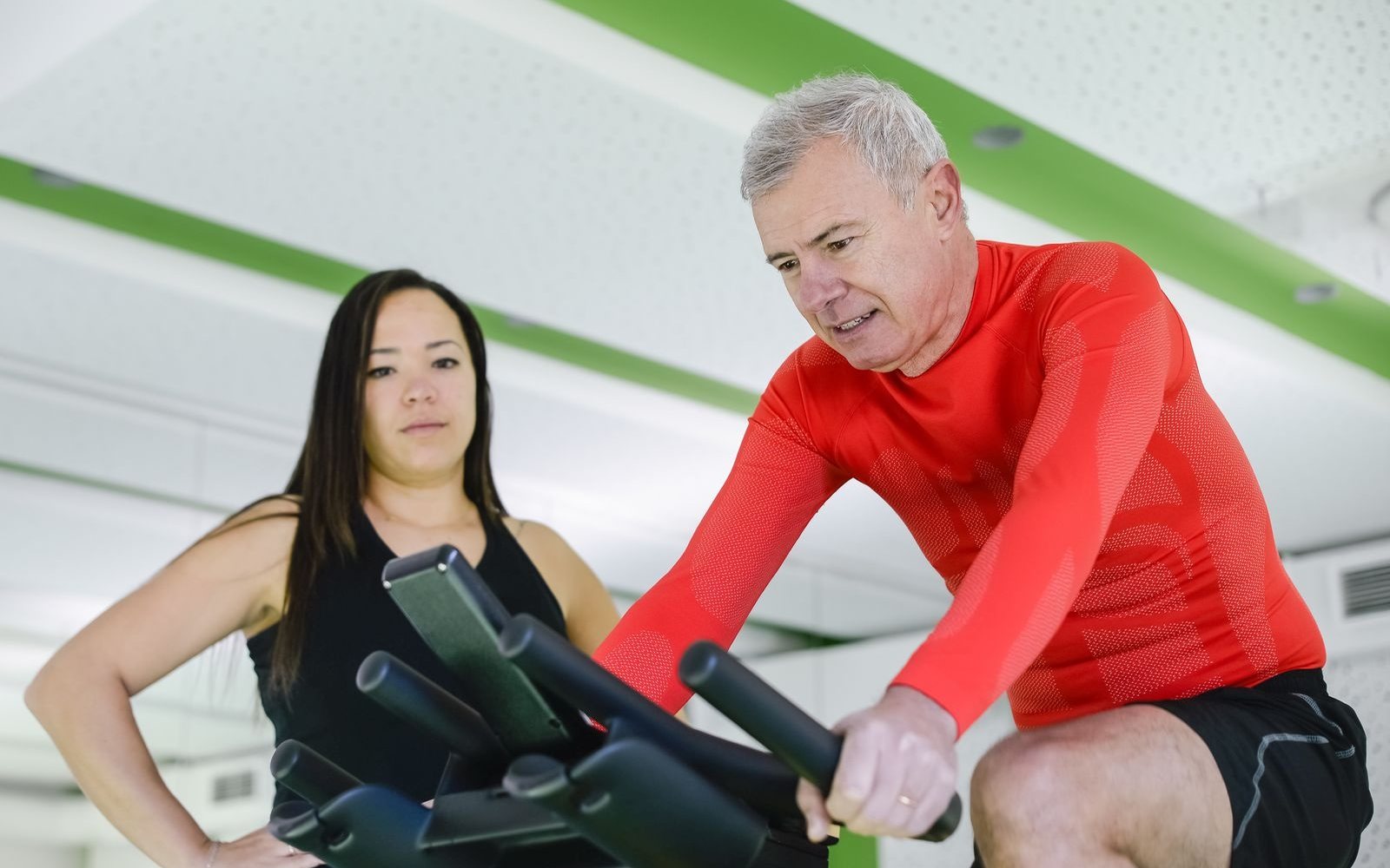 Older man pedaling a stationary bike while a trainer watches.