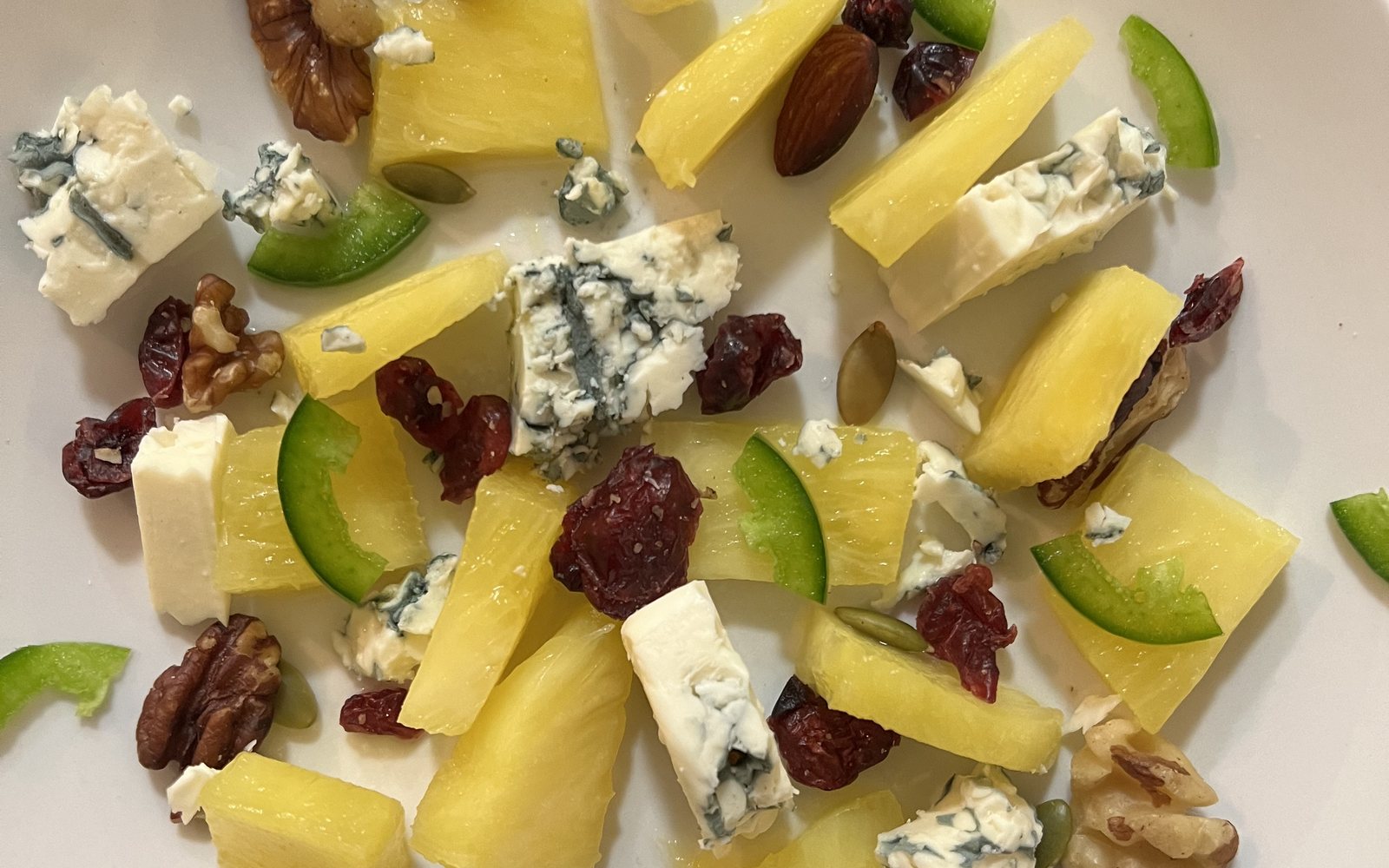 Pineapple, blue cheese, dried cranberries, and mixed nuts scattered on a white plate.