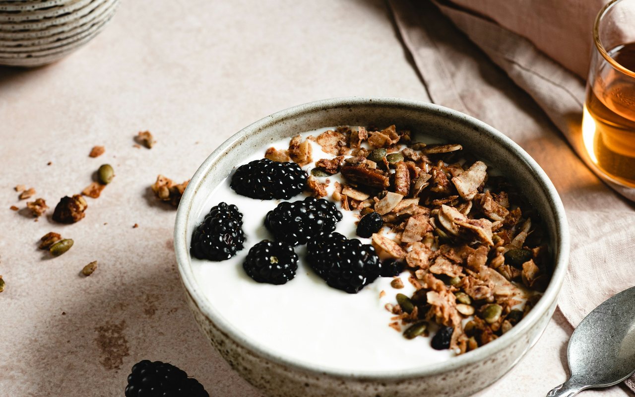 Greek yogurt topped with blackberries, granola, pumpkin seeds, and pecans in a ceramic bowl.