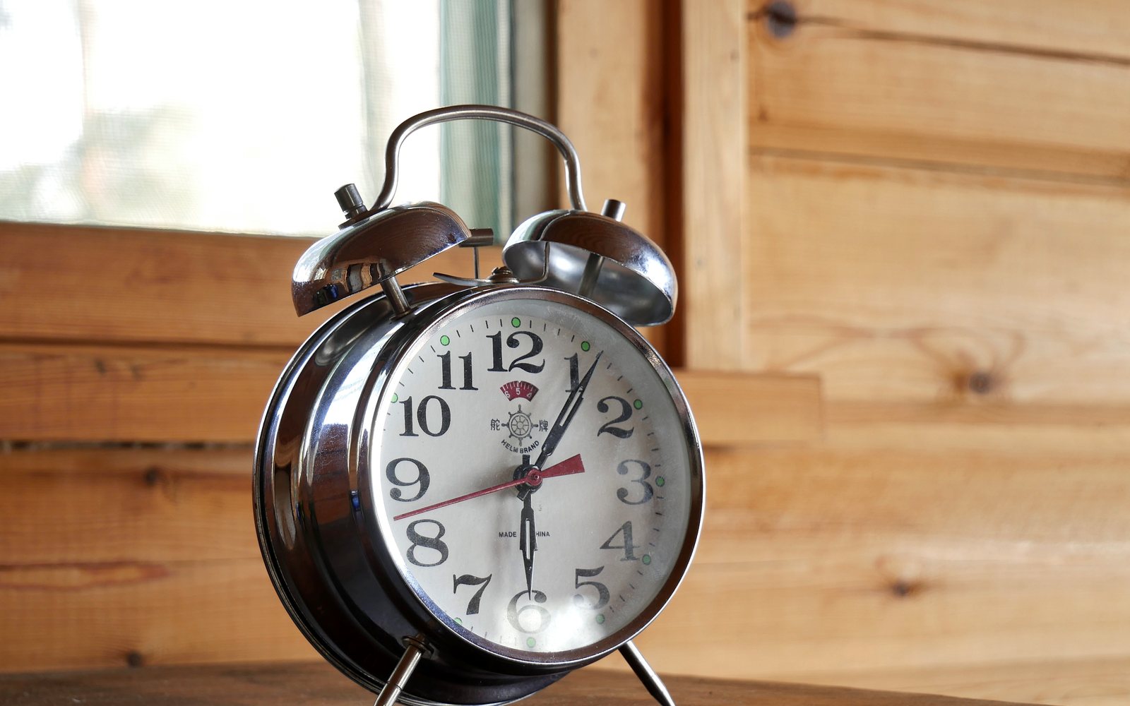 Chrome twin-bell alarm clock sits on a wooden surface beside a bright window.