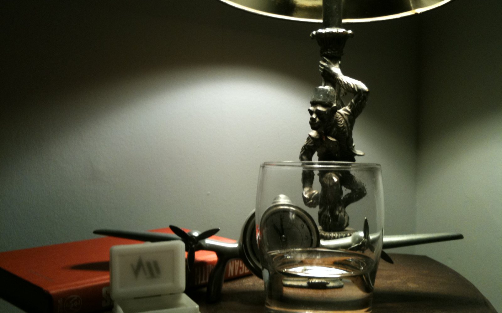 Dimly lit table with a clock, drinking glass, lamp, and model airplane.