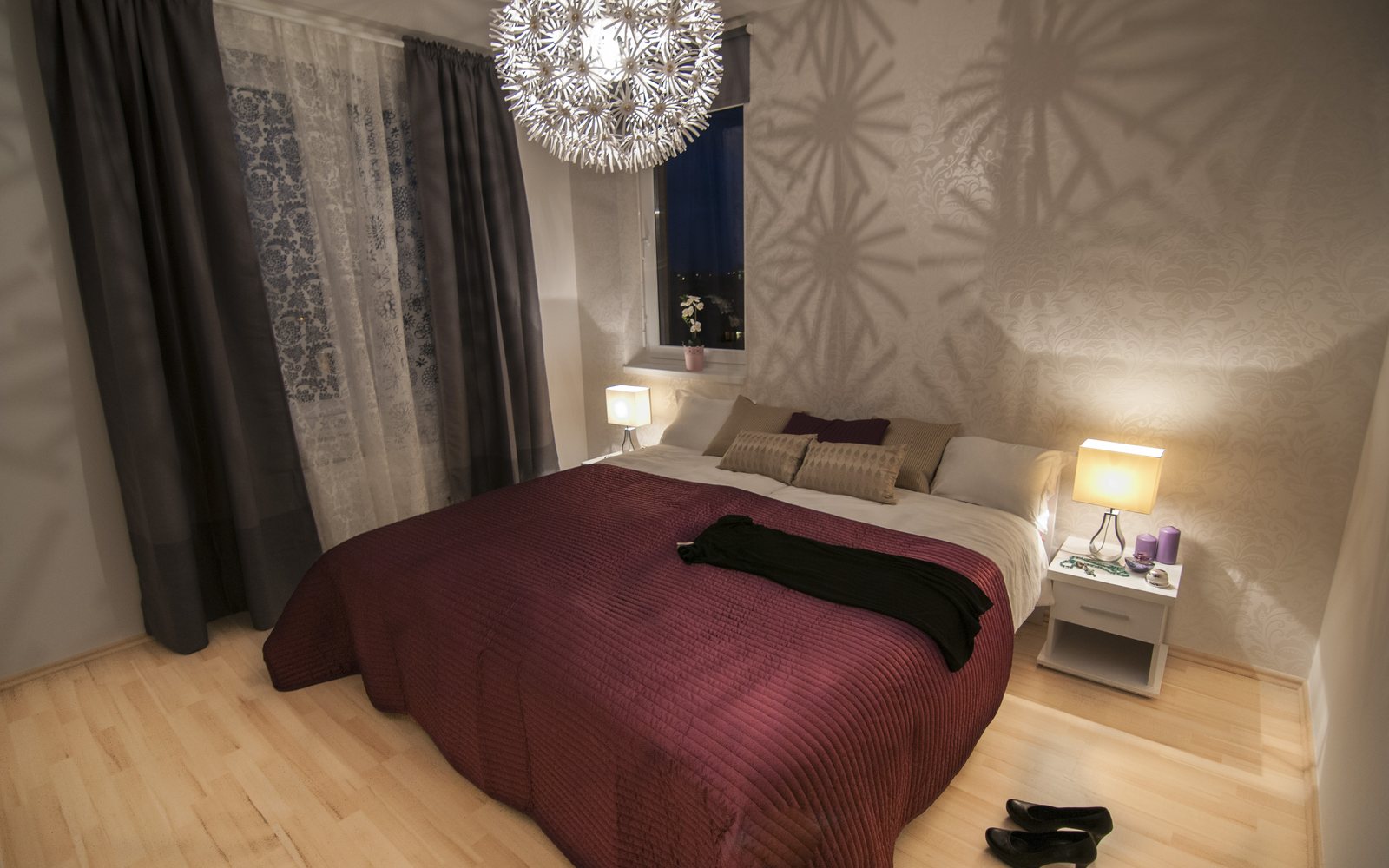 Made bed with a burgundy cover, bedside lamps, and dark curtains in a tidy bedroom.