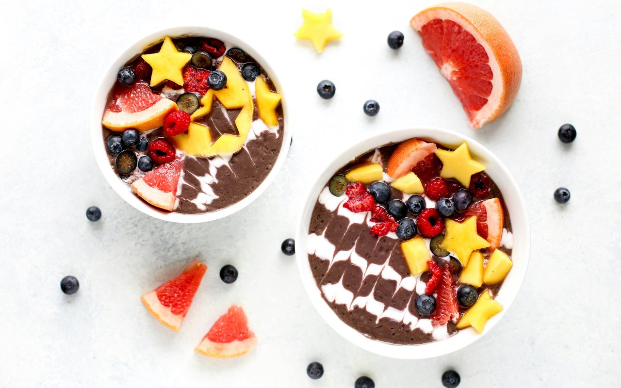 Two chocolate smoothie bowls topped with blueberries, raspberries, grapefruit, and mango stars on a white surface.