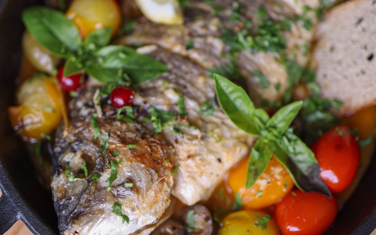 Whole baked sea bass surrounded by red and yellow cherry tomatoes and fresh herbs