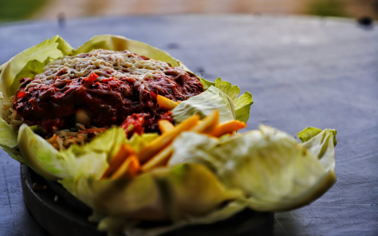 A lettuce-lined bowl filled with dark red sauced meat, shredded vegetables, and melted cheese.