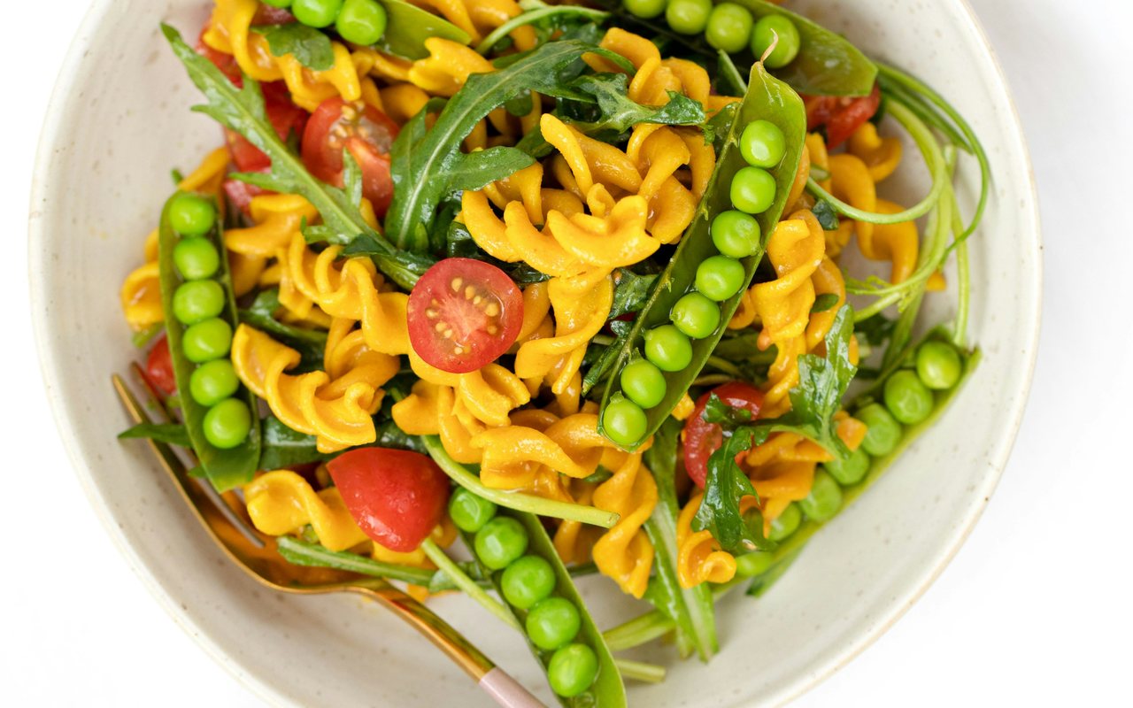 A bowl of orange lentil pasta with pea pods, cherry tomatoes, leafy greens, and summer vegetables.