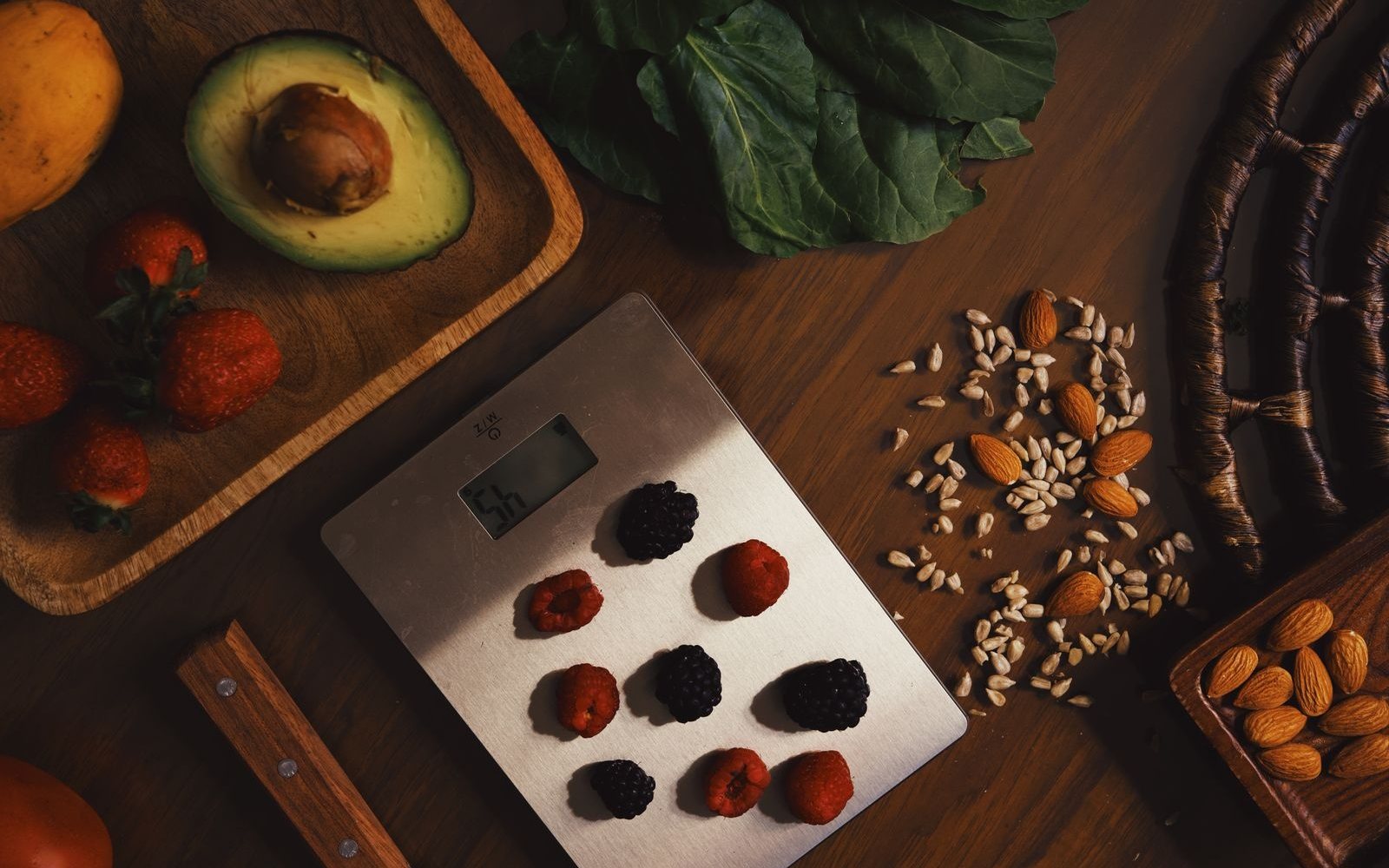 Berries on a kitchen scale surrounded by avocado, leafy greens, nuts, and seeds.