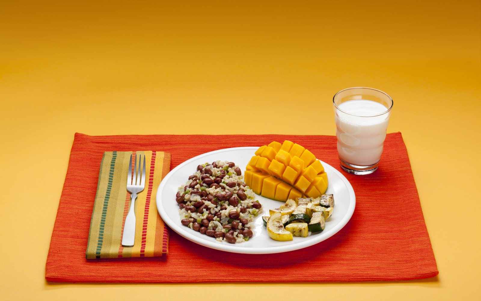 Plate of beans and rice, mango, and cooked vegetables beside a glass of milk.