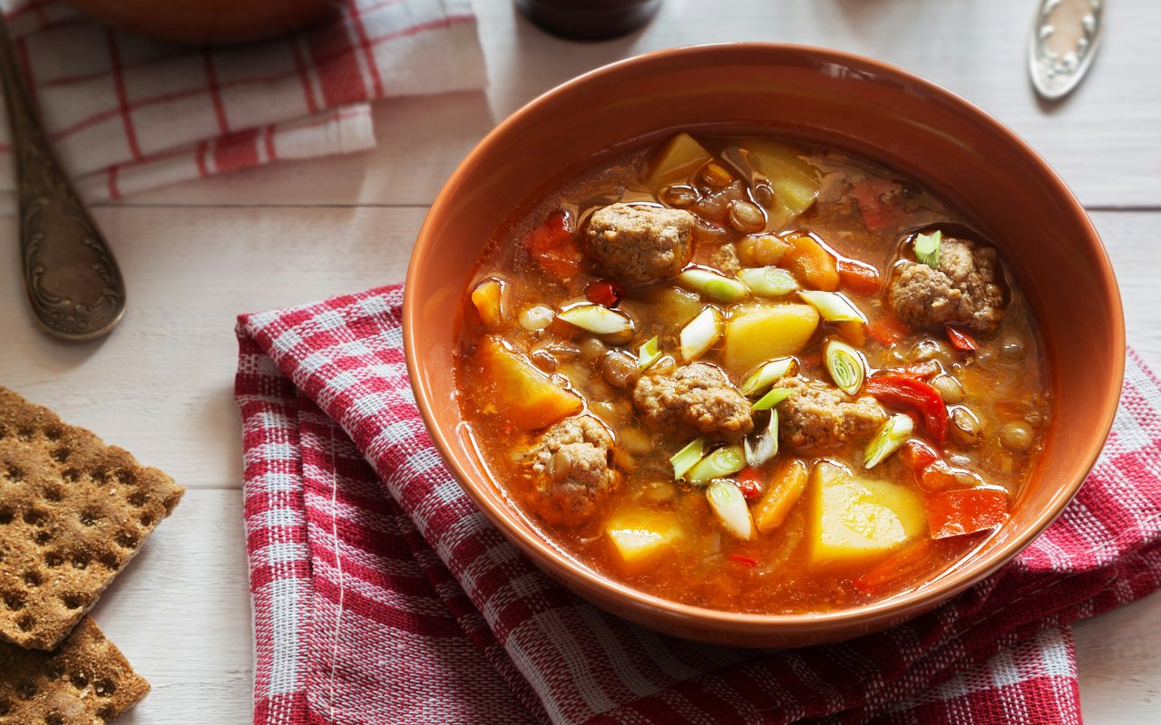 A terracotta bowl of lentil soup with meatballs, potatoes, carrots, red pepper, and scallions.